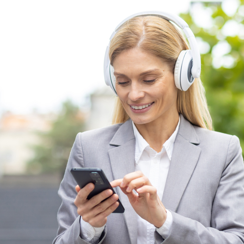 A white blonde woman wearing a gray blazer and headphones, listening to a podcast on her phone