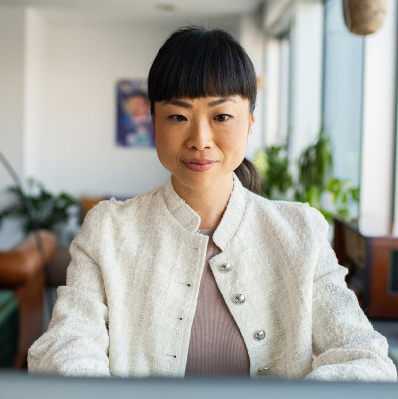 An asian woman wearing a white jacket seated in front of a laptop computer and looking intently at the screen