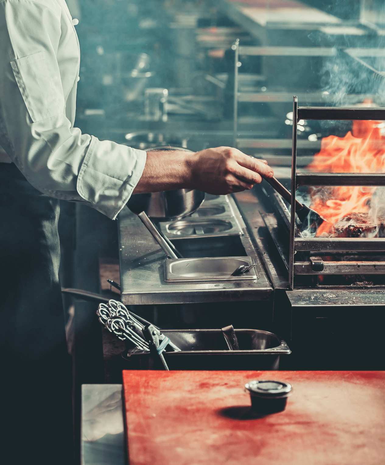 A chef preparing steak in a kitchen with flames shooting up from the grill.